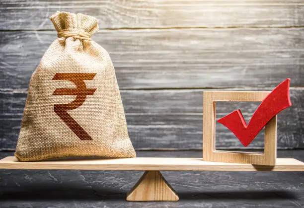 Photo of Red vote tick and a indian rupee money bag on scales. Estimating cost of making a decision and consequences in the future. Corruption risks. Concept of lobbying for decisions and laws.