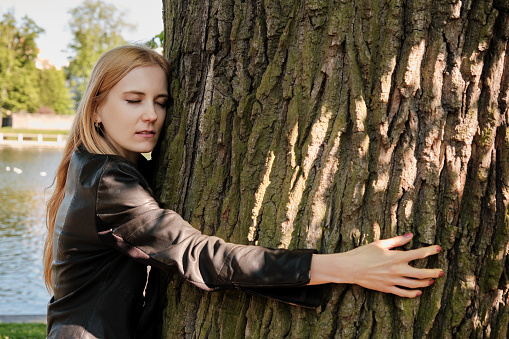 Pretty woman with long blond hair, wearing a leather jacket, hugging a giant oak tree trunk. Ecology concept.