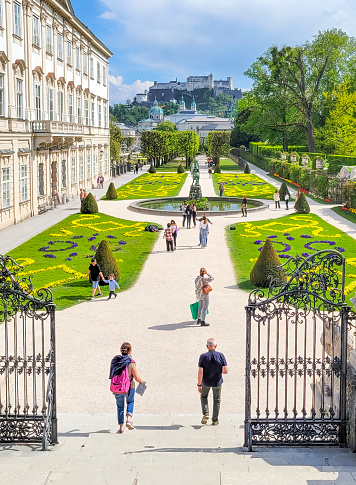 Mirabell gardens in Salzburg, Austria.  There are tourists walking through the formal box hedged gardens. It is a wide angle and also shows the fortress on the hilltop behind the gardens and the entrance gates in the foreground. The image was taken in May 2022.