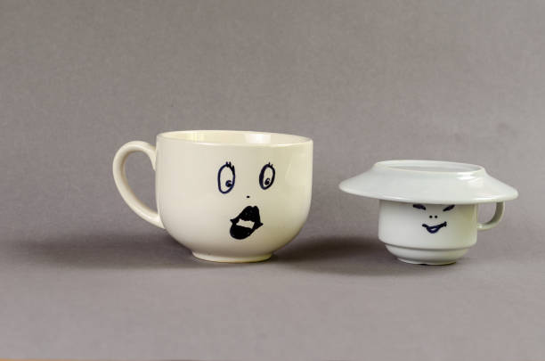istock Two ceramic cups against gray background. Large cup with a drawn surprised woman's face and puffy lips. Small cup with a man's face and a saucer instead of a hat. Flirt, love, relationship concept. 1405231766
