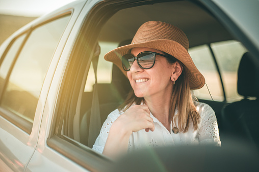 Smiling young woman in straw hat and sunglasses looking out the car window