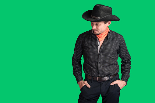 Cowboy in a black shirt looks to the side and to the bottom. An unshaven serious man in a hat, highlighted on a green background. Hands in pockets. Western American Culture