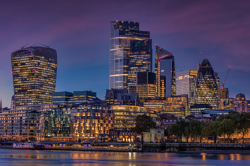 Twilight view of the illuminated skyscrapers of the City of London reflecting in the Thames River in the foreground.