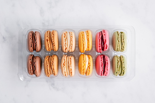 Overhead view of twelve rainbow coloured macaroon cakes in a plastic container on a minimal white marbled background
