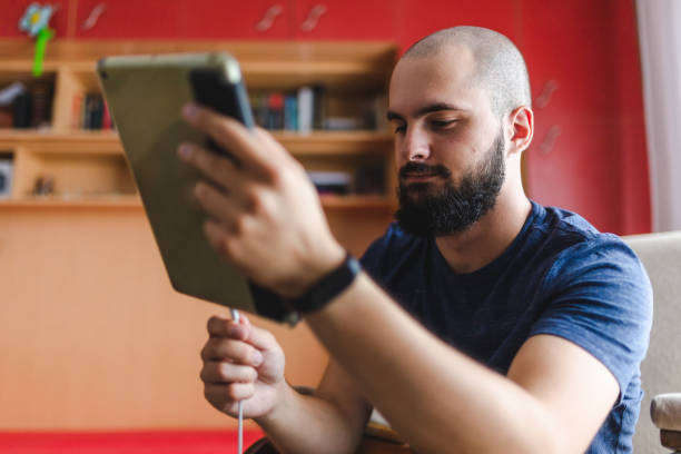 istock Close-up of a bald young man with a beard plugging in his iPad digital tablet to charge while relaxing in his room 1405252676