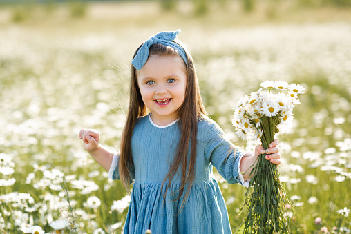 Smiling cute baby girl 3-4 year old wear stylish dress holding flower in blooming meadow outdoor. Childhood. Summer season.