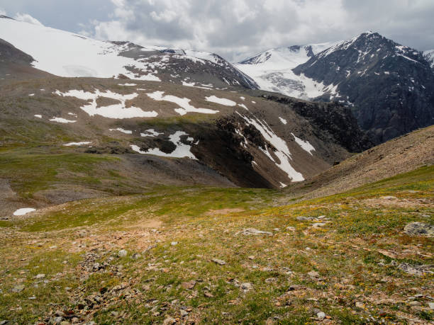 istock Summer view from sunlitgreen rocky slope to a narrow mountain gorge under cloudy sky. Colorful landscape with large snow mountains at changeable weather. 1405255607