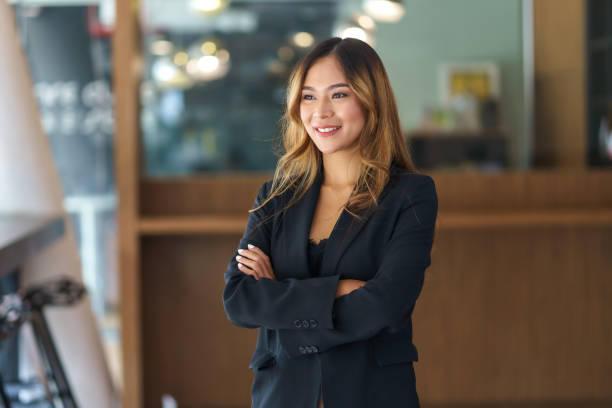 hermosa y sonriente mujer de negocios asiática de pie con los brazos cruzados en la oficina mirando por la ventana. - mirar hacia el otro lado fotografías e imágenes de stock