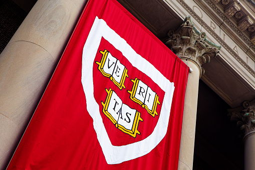 Cambridge, Massachusetts, USA - June 24, 2022: Close-up of the Harvard University logo on a red banner hanging from a building. The logo is a shield with the Latin motto 