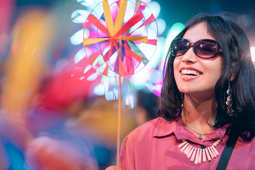 An Asian/Indian, happy, beautiful young woman enjoys and plays with colorful pinwheel toys in a fair in this waist-up image with copy space and selective focus. She wears sunglasses, a necklace, and a pink top. The Ferris wheel illuminates in the crowded background.