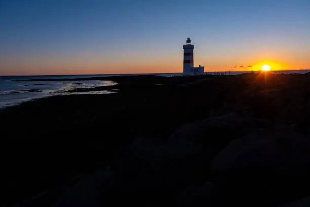 Photo of Beautiful view of white and red colored old lighthouse Garður on the coast of Suðurnesjabær on Reykjanes peninsula, Iceland with beach during midnight sun