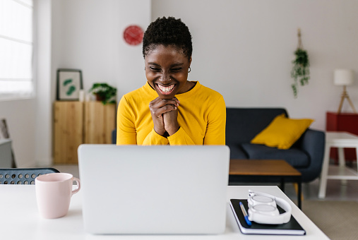 Excited young african woman celebrating success looking at laptop screen - Millennial female reading good news on computer while sitting at workplace - Winning and celebration concept
