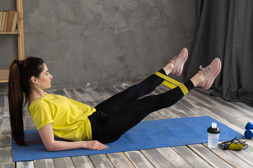 happy woman exercising with resistance band lying on mat at home. athlete swings her legs with sport fitness rubber bands. sport, fitness and people concept
