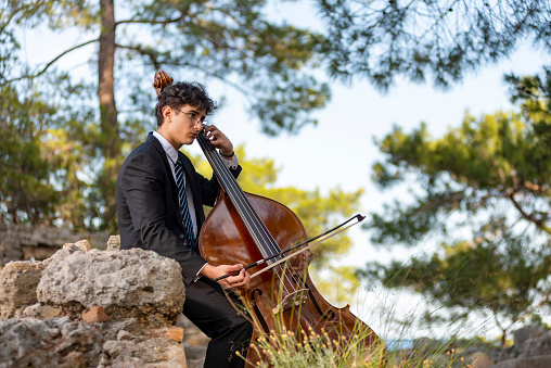 Young musician in a black suit plays double bass in nature. He sits on a rock and makes music in the summer.