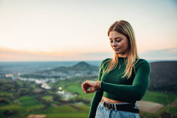 Young blonde millennial generation Girl standing outdoors in the nature during sunset looking down to her smart watch, checking messages, news and fitness data of his walk - hike. South German Mountain Range in the background. Backlit from Dusk - Sunset Sun. Real People Millennial Generation Outdoors Modern Technology Fitness Scene.