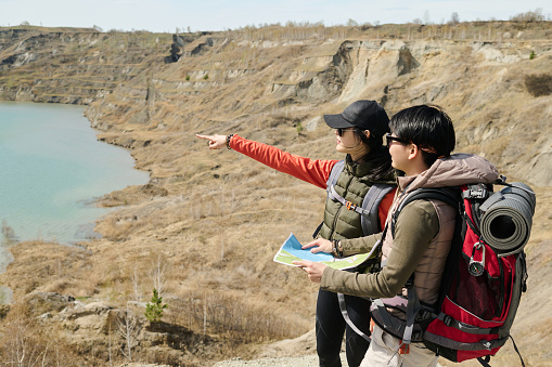 Two young Asian women wearing backpacks hiking in mountains on sunny spring day reading paper map and choosing direction