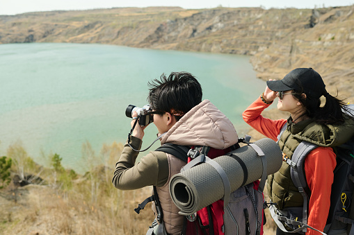 Two active young Asian female tourists wearing backpacks hiking around quarry lake enjoying view and taking photos on camera