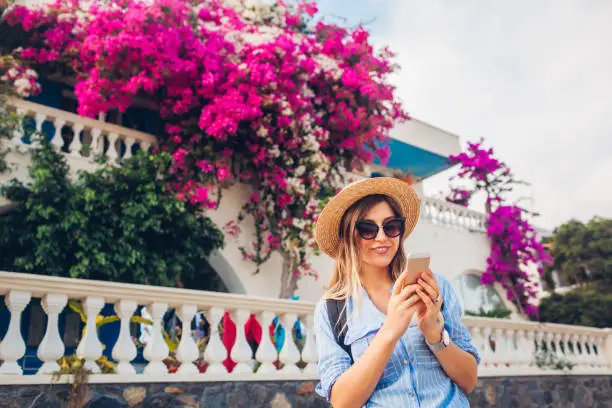 Woman texting on smartphone on street with blooming bougainvillea flowers. Happy woman wearing hat and backpack. Summer nature among city architecture