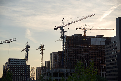 Dark silhouettes of tower cranes and high-rise buildings under construction at construction site at sunset. Blue yellow evening sky