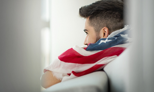 Closeup side view of a young man at home looking through a window and having American flag on his back on Fourth of July.