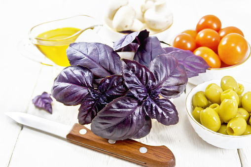 Fresh purple basil in a mortar, olives, tomatoes and champignons in bowls, vegetable oil in gravy boat and a knife on white wooden board background