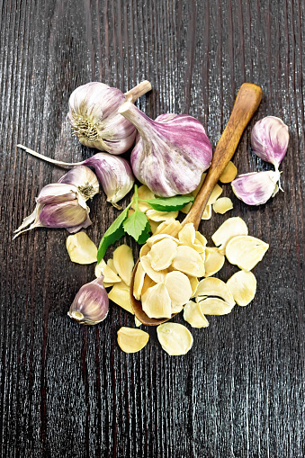 Dried garlic slices in a spoon and on the table, fresh garlic, basil on dark wooden board background from above