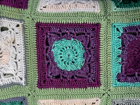 Horizontal high angle closeup photo of a section of a handmade crocheted knee rug with green, cream, turquoise and purple coloured squares.
