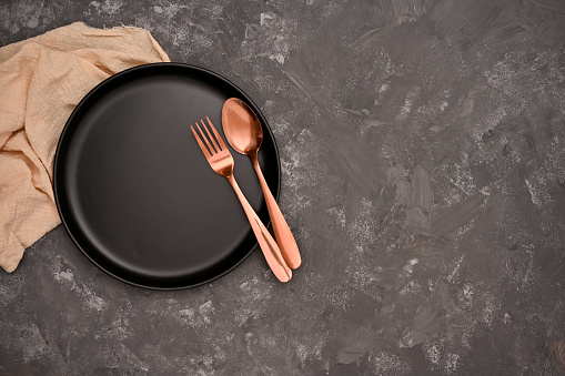 Luxury restaurant dining table setting with empty black ceramic plate, golden fork and spoon, napkin on black background. top view