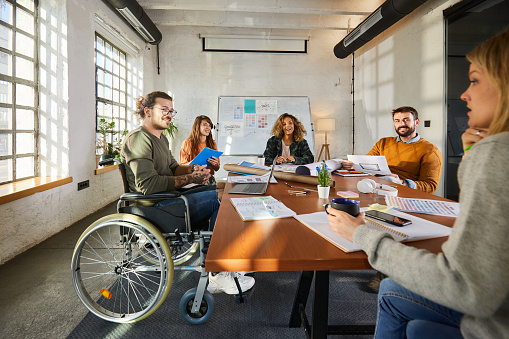 Happy creative man in wheelchair communicating with his colleagues during a meeting at casual office.