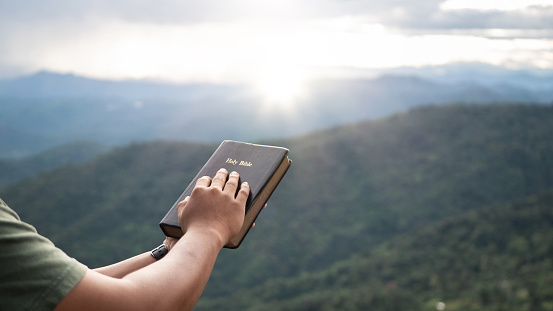 man praying on the holy bible in a field during beautiful sunset.male sitting with closed eyes with the Bible in his hands, Concept for faith, spirituality, and religion.