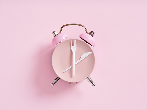 Alarm clock with plate and cutlery on pink background. Intermittent fasting, lunchtime, weight loss, meal plan or diet concept.