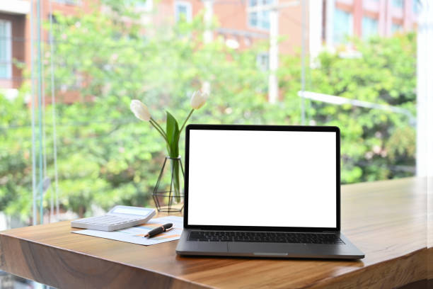 istock Mockup laptop computer with white empty screen on wooden table near large office window. 1405357585