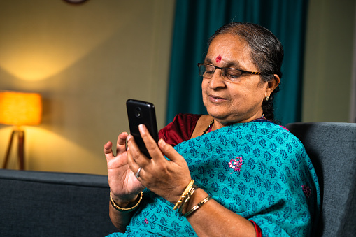 Senior woman busy using mobile phone while sitting on sofa at home - concepts of technology, relaxation and social media.
