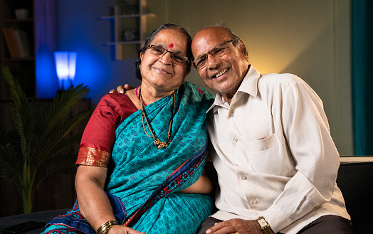 portrait shot of Happy senior couple embracing each other by looking camera at home- concept of relationship, happiness and family bonding.