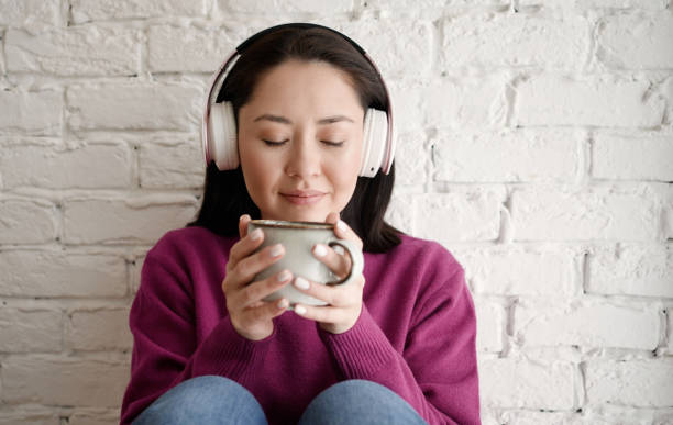 jeune femme asiatique multiethnique rêveuse au casque tenant une tasse de thé boire une boisson au café du matin tout en étant assise à la maison dans la chambre à coucher - the thinker audio photos et images de collection