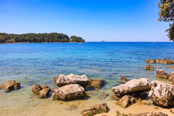 Big rocks on sandy beach and blue seascape with land in the distance, Rovinj riviera under the clear sky