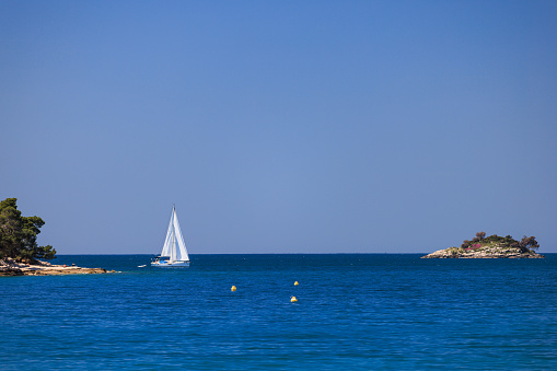 White sailboat floating on the sea surface by the rocky coastline with littoral vegetation at remote location, clear blue sky and island in the distance