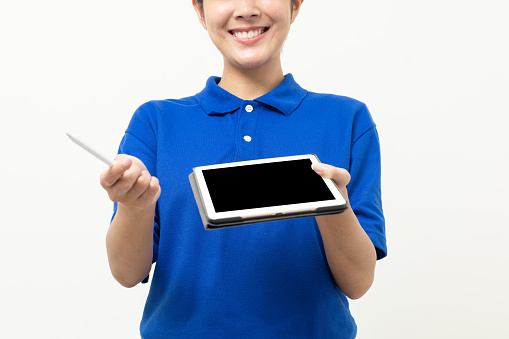 Happy delivery asian woman in blue uniform standing holding digital tablet signing to receive for customer. Female worker checking product with tablet standing on isolated white background.
