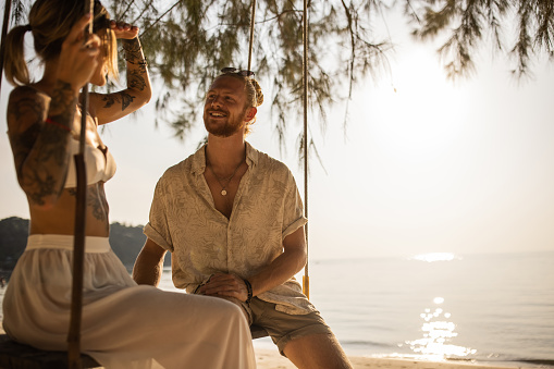 Young Caucasian couple in love enjoy the sunset on the beach, while swinging on the rope swing, during their vacation