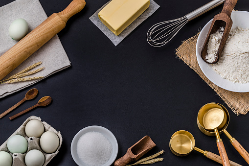 Top view of various bakery  ingredients and items such as eggs, butter, flour, sugar, rolling pin, measure cups, hand whisk and serving scoop disposed all around the image leaving a useful copy space at the center on black background.