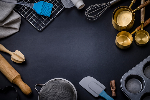 Top view of various pastry  utensils a rolling pin, measure cups, hand whisk, colander, baking mold and serving scoop disposed at the top of the image leaving a useful copy space at the lower side on black background.