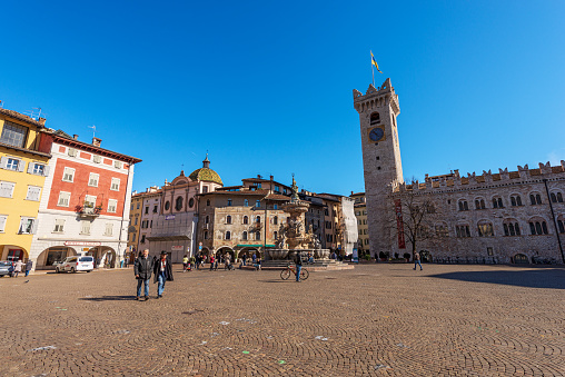 Trento, Trentino Alto Adige, Italy - March 8th, 2020: Piazza del Duomo, Cathedral square in Trento city with the Neptune fountain, Civic tower (Torre Civica), Praetorian Palace (Palazzo Pretorio or Palazzo Vescovile) and the frescoed houses Cazuffi Rella, XVI century. A group of tourists and locals stroll around the large square or relax sitting at one of the bars on a sunny late winter day.