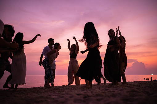 Group of multiracial female and male friends, enjoy the sunset on the beach, while dancing and playing guitar