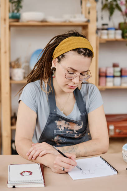 istock Young creative female potter making sketch of earthenware product in notepad 1405392466