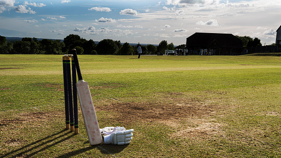 Wide angle shot of a cricket bat leaning on stumps with gloves on ground,  shot in late afternoon sun
