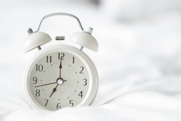istock Alarm clock on white bad for wak up time with light from window, selective focus 1405395811