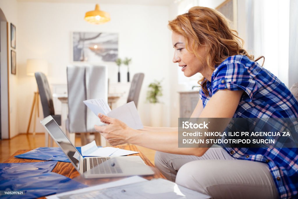 Woman paying bills and working online stock photo Smiling glad woman paying bills online at home while sitting in living room, doing paper work and using laptop computer at her workplace wearing casual clothes Financial Loan Stock Photo