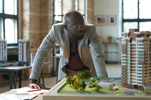 African serious architect in eyeglasses looking at construction model of new residential area during his work at office