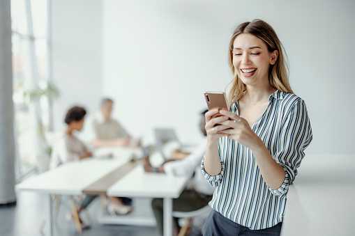 Young beautiful female entrepreneur with smartphone and confident expression as other workers hold meeting in background. Portrait of caucasian businesswoman in meeting room and copy space