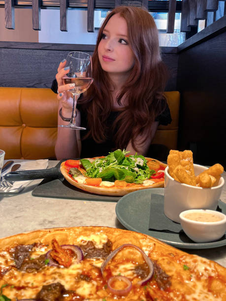 Close-up image of beautiful young woman with red hair in restaurant setting, holding glass of rose wine whilst sat at restaurant table, meal of pizza and salad with polenta chips, profile view stock photo